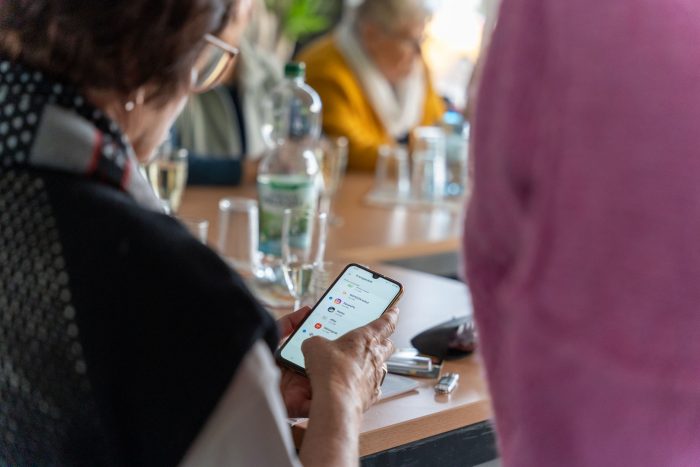 Eine Person, die ein Smartphone in einer Besprechung hält, während andere Teilnehmer im Hintergrund sichtbar sind.