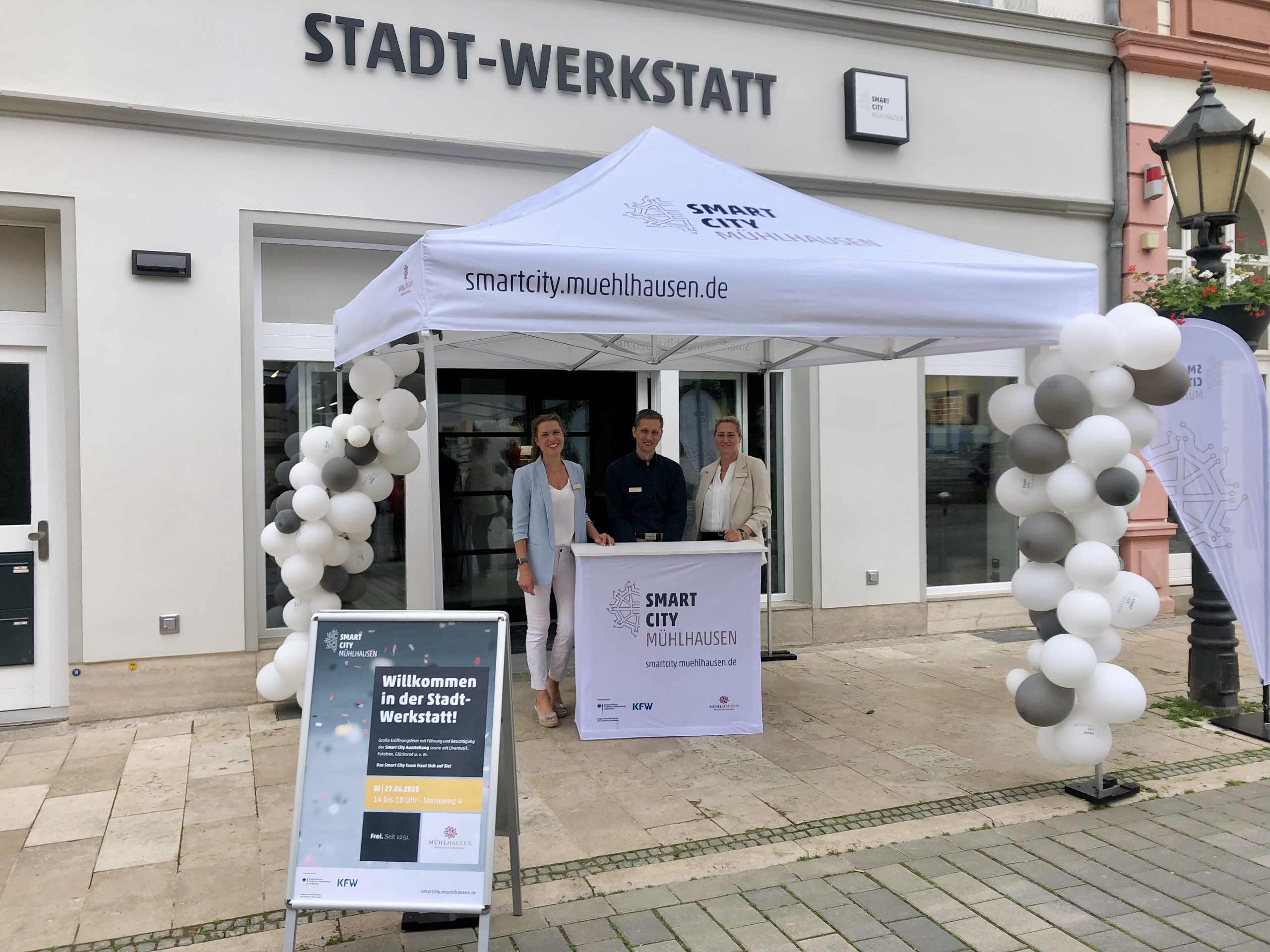 Drei Personen stehen unter einem Zelt der Stadt-Werkstatt in Mühlhausen, umgeben von Luftballons.