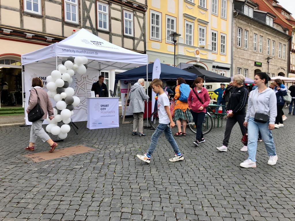 Ein Marktstand mit Informationen zur Smart City, umgeben von Besuchern auf einem Platz.