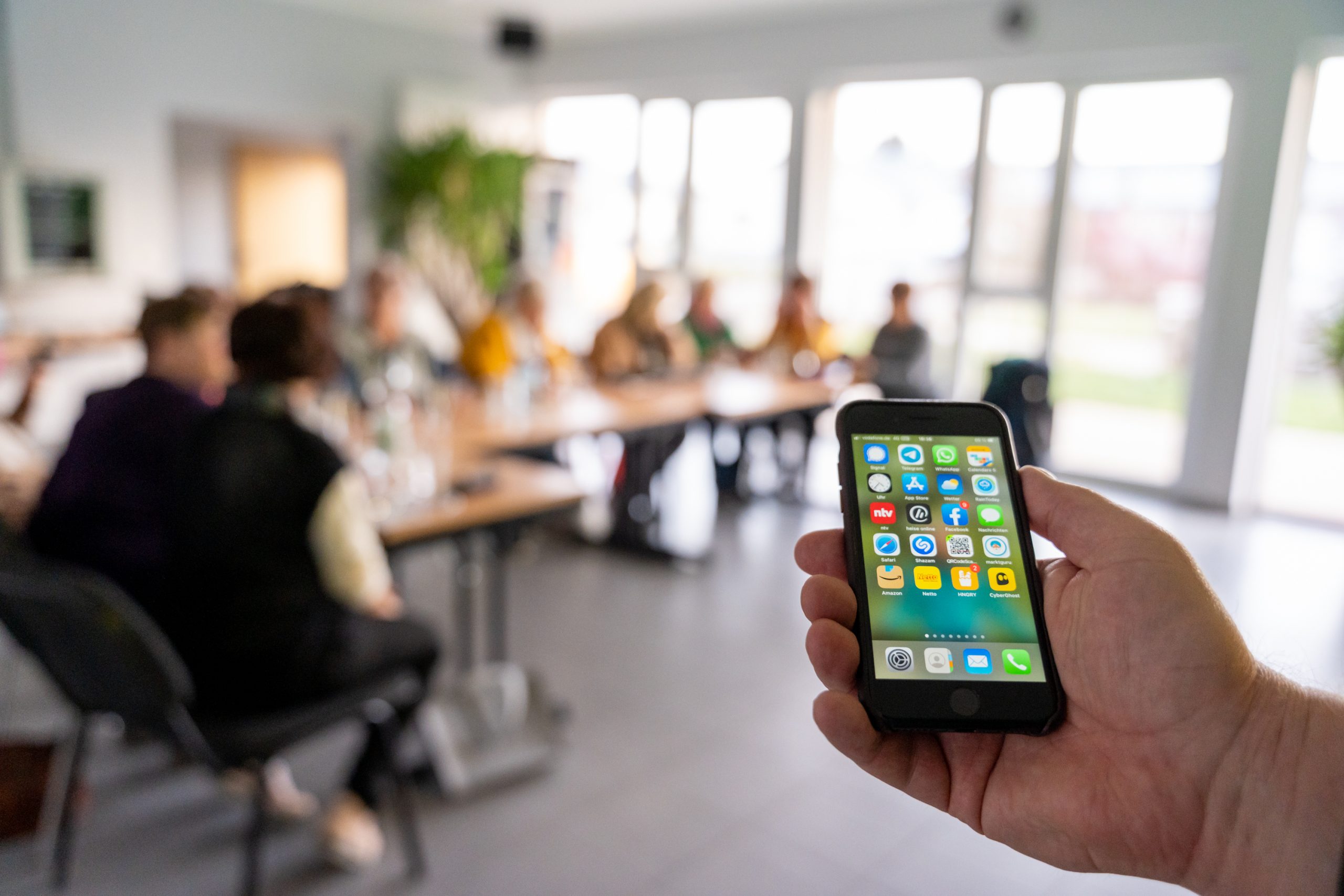 Ein Smartphone mit verschiedenen App-Symbolen wird in einer Besprechung gehalten, während Personen im Hintergrund sitzen.