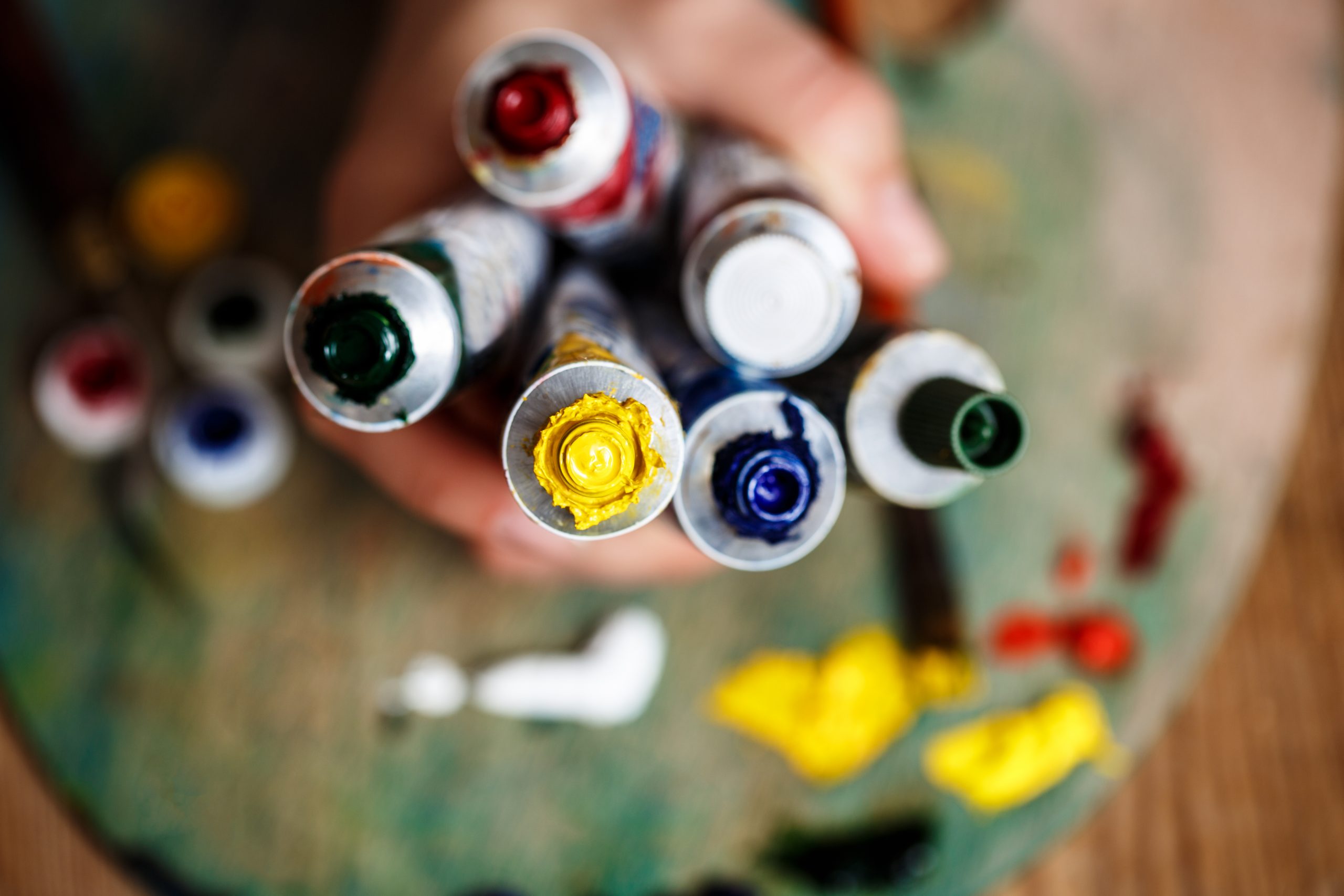 Eine Hand hält mehrere Tuben mit Acrylfarbe in verschiedenen Farben, darunter Gelb, Blau und Rot.