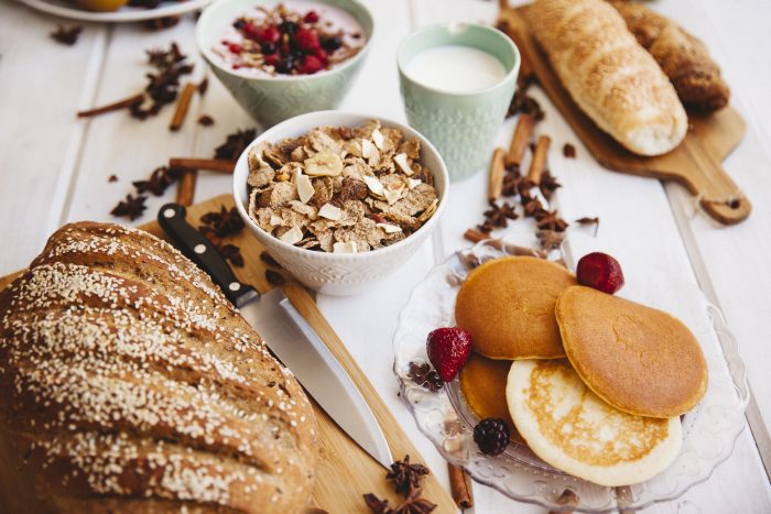 Ein Frühstückstisch mit Brot, Müsli, Joghurt und Pfannkuchen auf einem weißen Tisch.