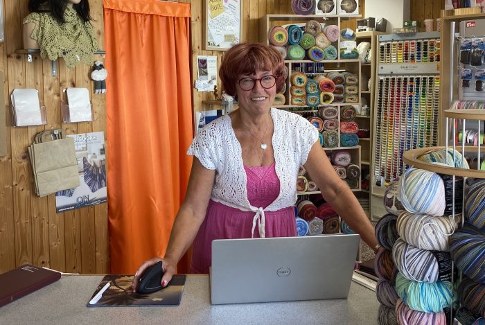 Eine Frau mit roten Haaren steht an einem Tisch mit einem Laptop in einem Wollgeschäft.