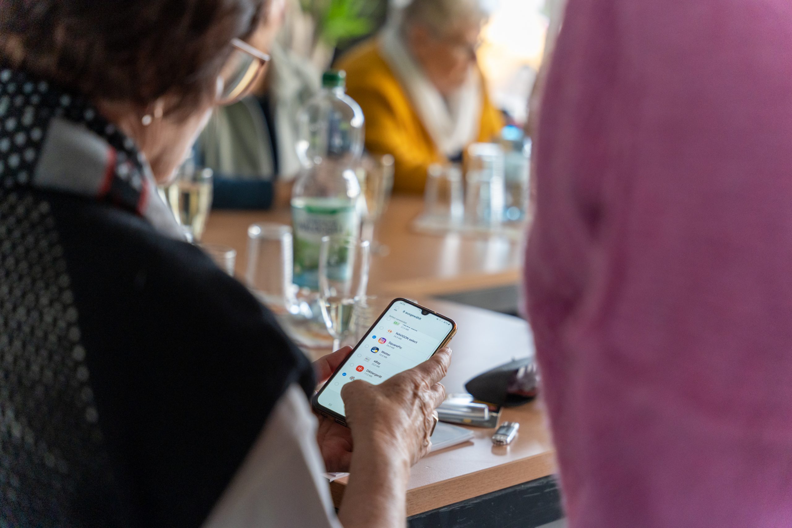 Eine Person hält ein Smartphone und betrachtet eine App während einer Besprechung mit anderen Personen im Hintergrund.