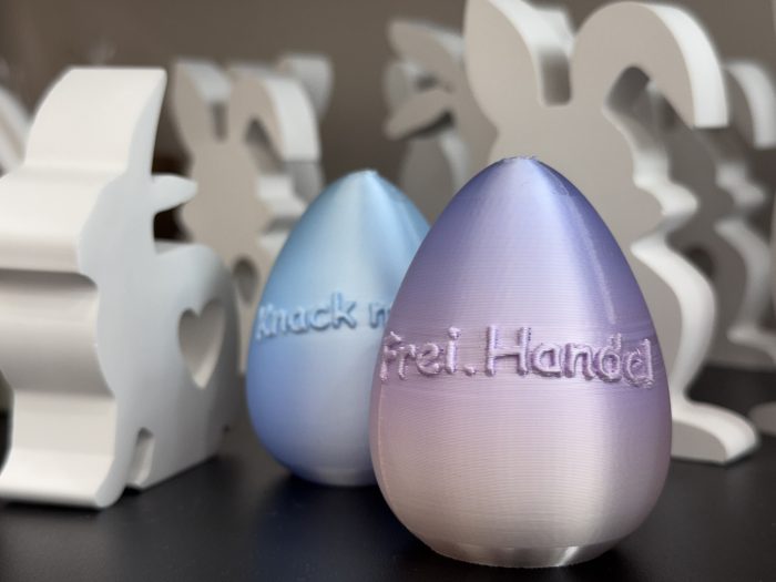 Zwei 3D-gedruckte Ostereier in Pastellfarben mit eingravierten Texten auf einem Tisch.