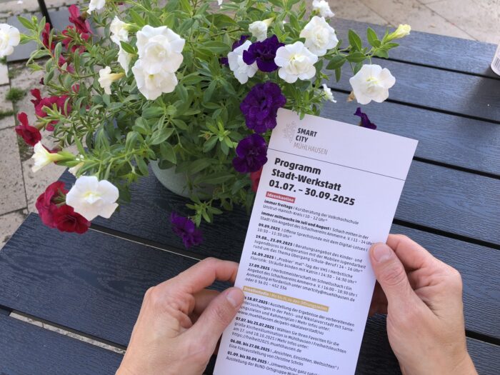Hand hält ein Informationsblatt über das Programm der Stadt-Werkstatt vom 01.07. bis 30.09.2025.