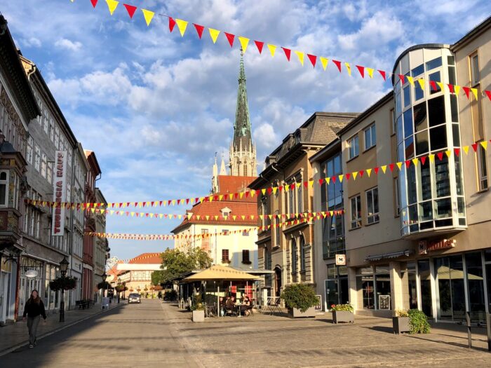 Blick auf eine Straße mit bunten Fahnen und einem Kirchturm im Hintergrund.
