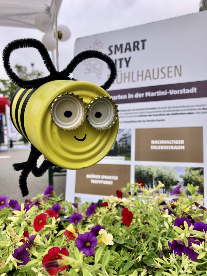 Biene aus einer gelben Dose mit Augen aus Flaschendeckeln, hängt an einem Stand.