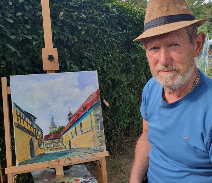 Ein Mann mit Bart sitzt neben einer Staffelei, auf der ein Landschaftsgemälde zu sehen ist.