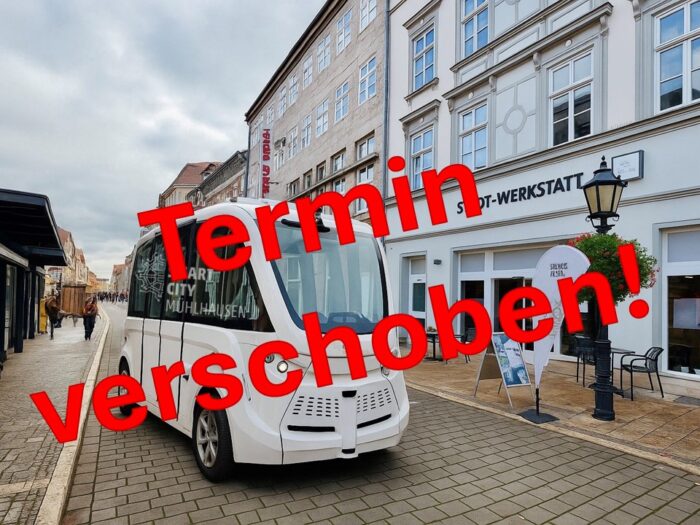 Weißer autonomer Shuttlebus mit dem Text 'Termin verschoben!' auf einem Stadtweg.