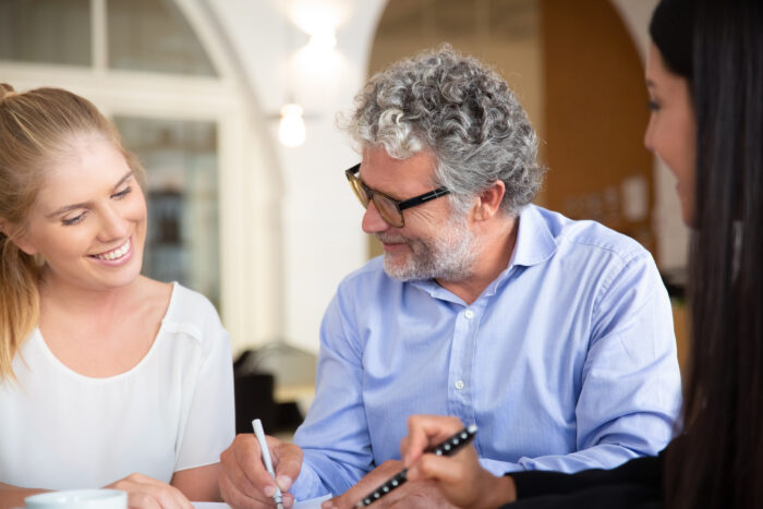 Drei Personen in einem Gespräch, eine Frau lächelt, ein Mann mit Brille spricht, eine andere Frau macht Notizen.
