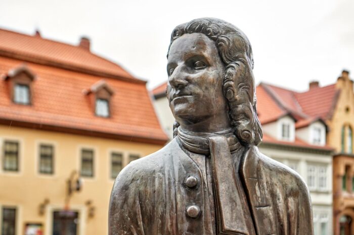 Bronzene Skulptur eines historisch gekleideten Mannes mit langer Lockenperücke, frontal sichtbar, Knöpfe und Schal der Weste detailreich, Hintergrund mit hellen Gebäuden und roten Ziegeldächern