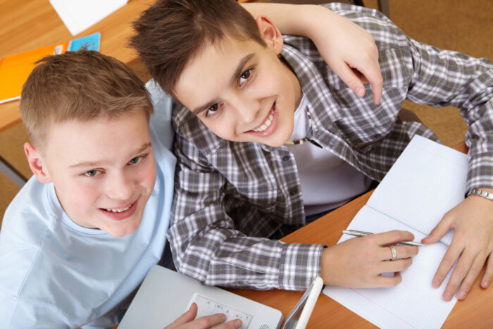 Zwei Jungen sitzen an einem Schreibtisch und lächeln in die Kamera, einer hält einen Stift.