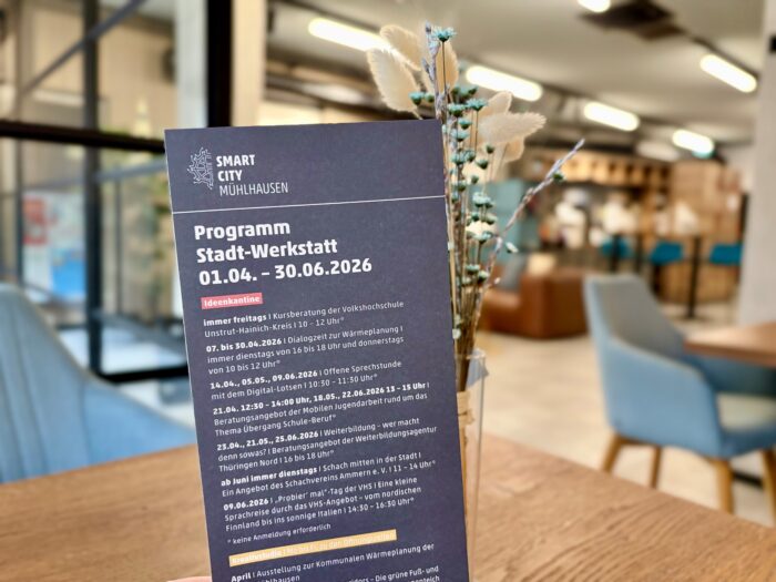 Dunkelblaues Poster mit dem Text Programm Stadt-Werkstatt 01.04. - 30.06.2026, umgeben von Trockenblumen in einer Vase, im modernen Innenraum mit Blick auf Sitzbereich.