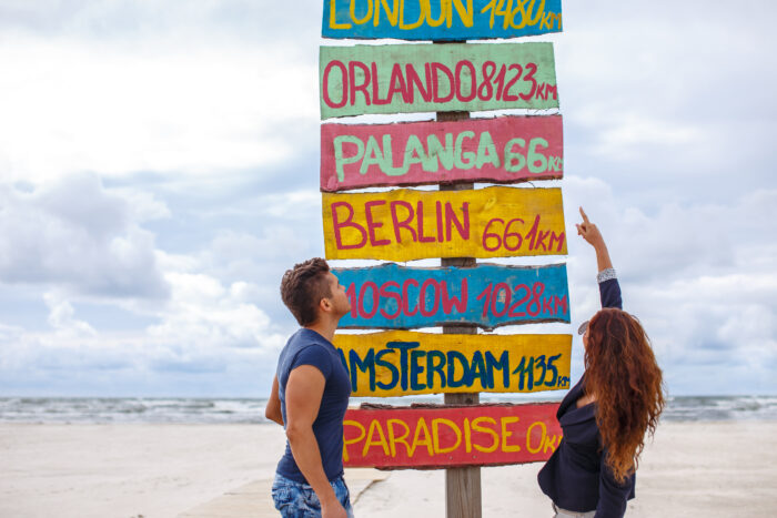 Vor einem Strand stehen zwei Personen neben einem bunten Wegweiser mit Entfernungen zu London, Orlando, Palanga, Berlin, Moskau und Amsterdam.