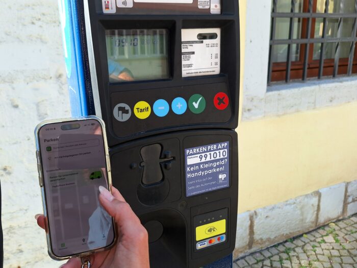 Vor einem schwarzen Parkautomaten hält eine Hand ein Smartphone mit der Parken-App, daneben sind Tarif-, Karten- und App-Hinweise zu sehen.