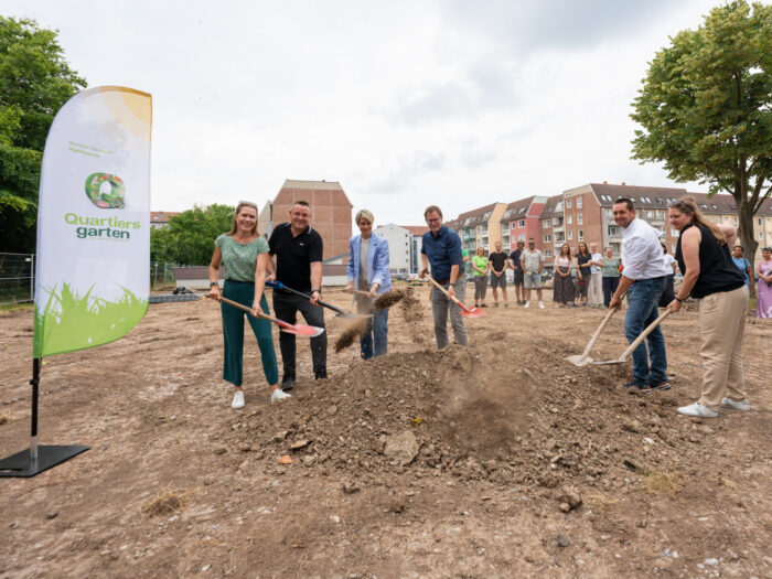 Menschen werfen bei einem Spatenstich Erde auf einem brachliegenden Gelände neben einem Quartiersgarten-Banner in einem Wohnviertel auf.